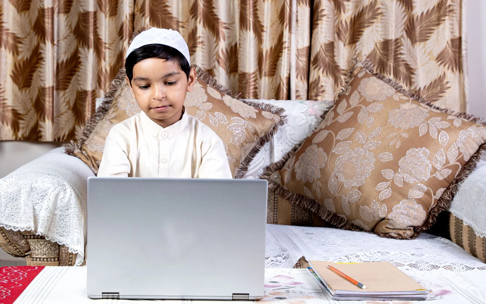 Young boy in a kufi learning Quran online on a laptop at home