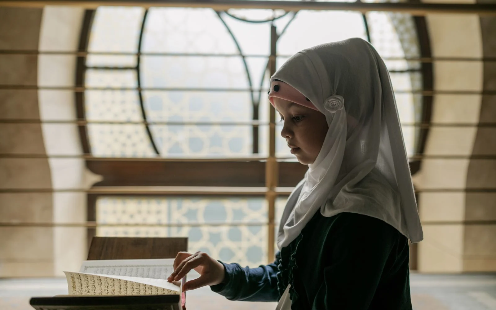 Girl in hijab reading the Quran from a wooden stand by a window